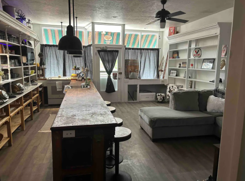 A cozy interior featuring a wooden bar, stools, a sectional sofa, and shelves with decor and natural light.